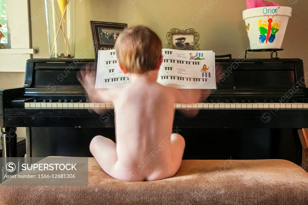 Two year old boy, sitting naked on a bench, playing an upright piano.