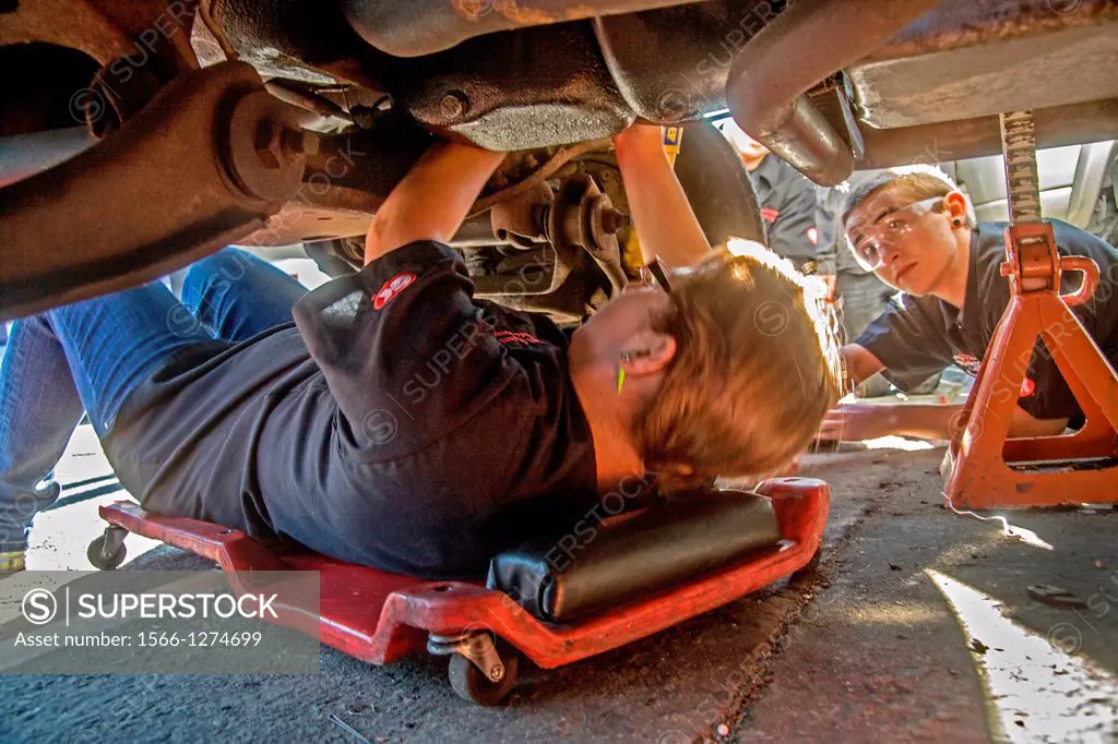 Wearing safety glasses a Caucasian teenage girl and boy work underneath a car engine in auto shop class in San Clemente, California, USA