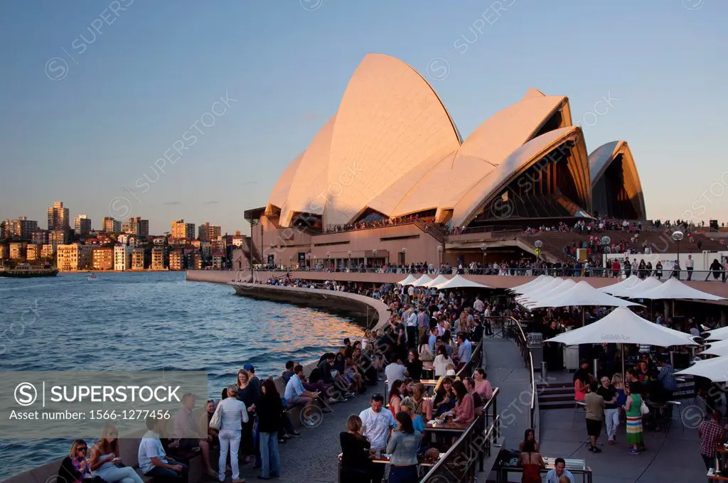 Last rays of the sun on Sydney Opera House, many people drinking and eating at restaurants and bars with outdoor seating. Australia, New South Wales, ...