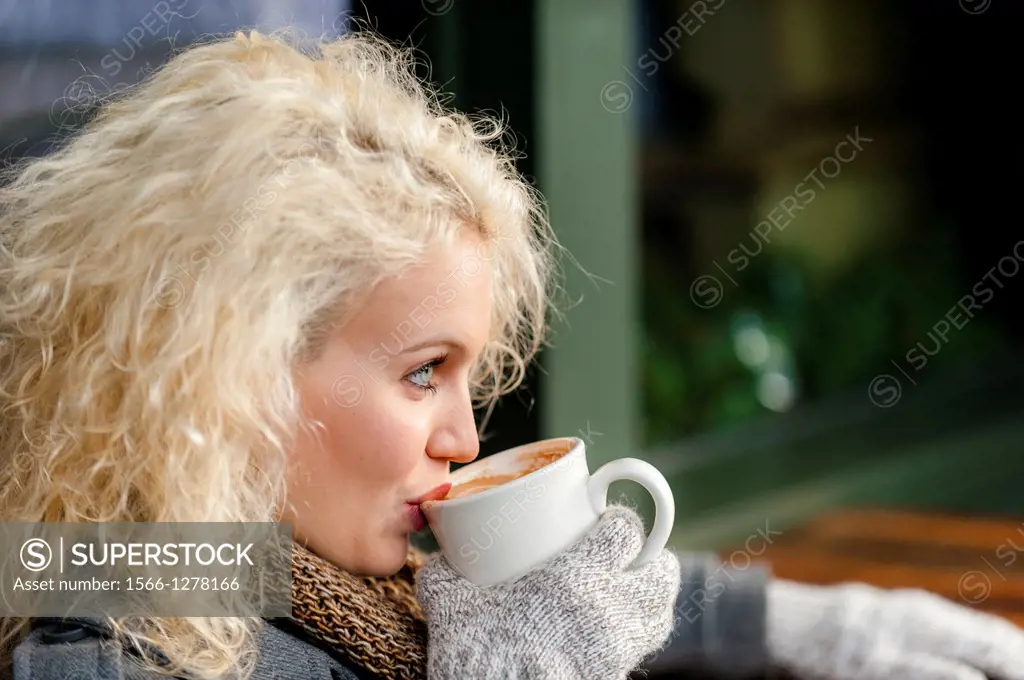 A 22 year old blond woman sitting at an outdoor cafe with a cup of coffee in the winter.