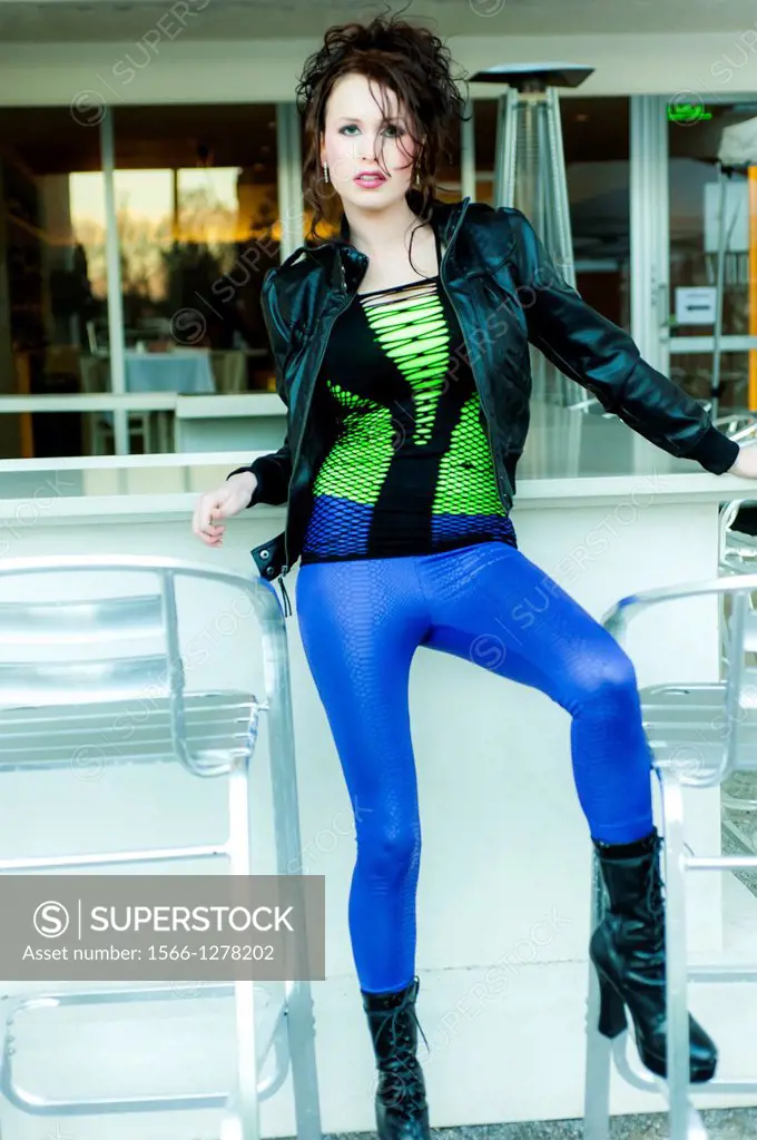 A 24 year old brunette woman in weaing blue leggings and a black jacket standing at an outdoor bar.