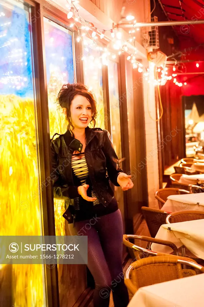A 24 year old brunette woman in weaing leggings and a black jacket standing at an outdoor cafe.