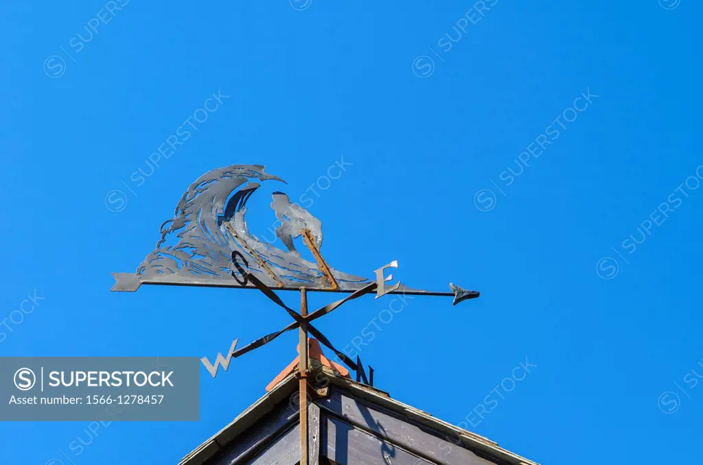 A surfer riding a wave weather vane on the gable of a roof against a blue sky.