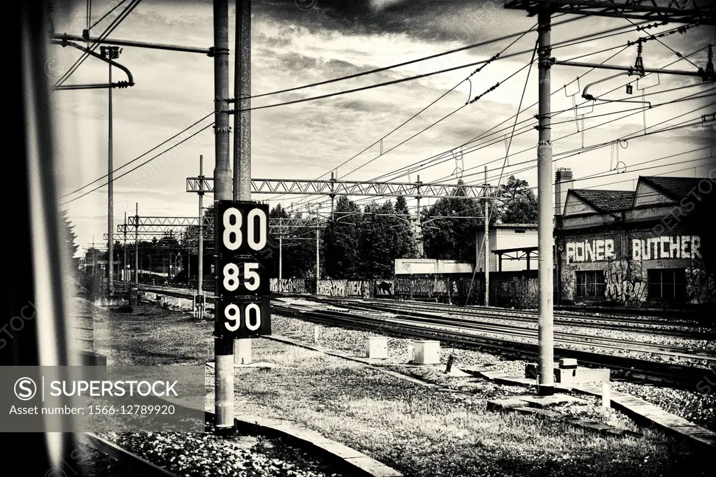 View from a train window, of the industrial area with industrial buildings, rail tracks and wires and a sign post with numbers, 80, 85, y 90. Milano, ...