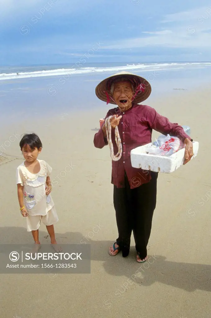 Hoi An; Beach; old Woman with a little Child; selling necklets; Traditional Costume; Woman wearing traditional conical Hat; Vietnam.