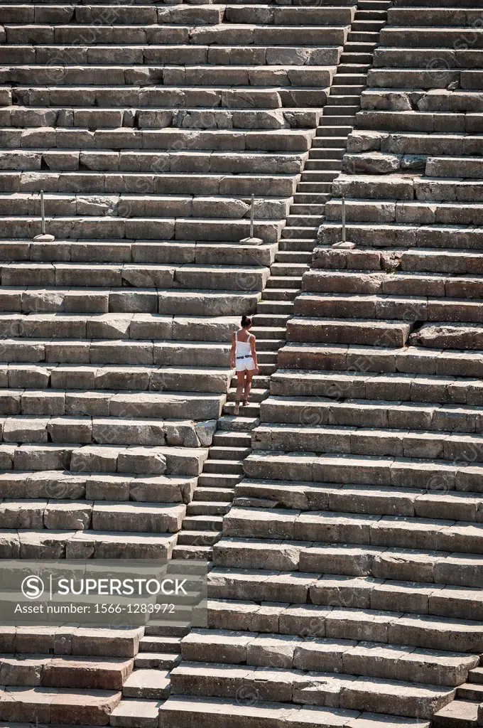 A woman climbs steps amongst the tierd seating of the Classical Greek theatre at Ancient Epidaurus, Argolid, Peloponnese, Greece.