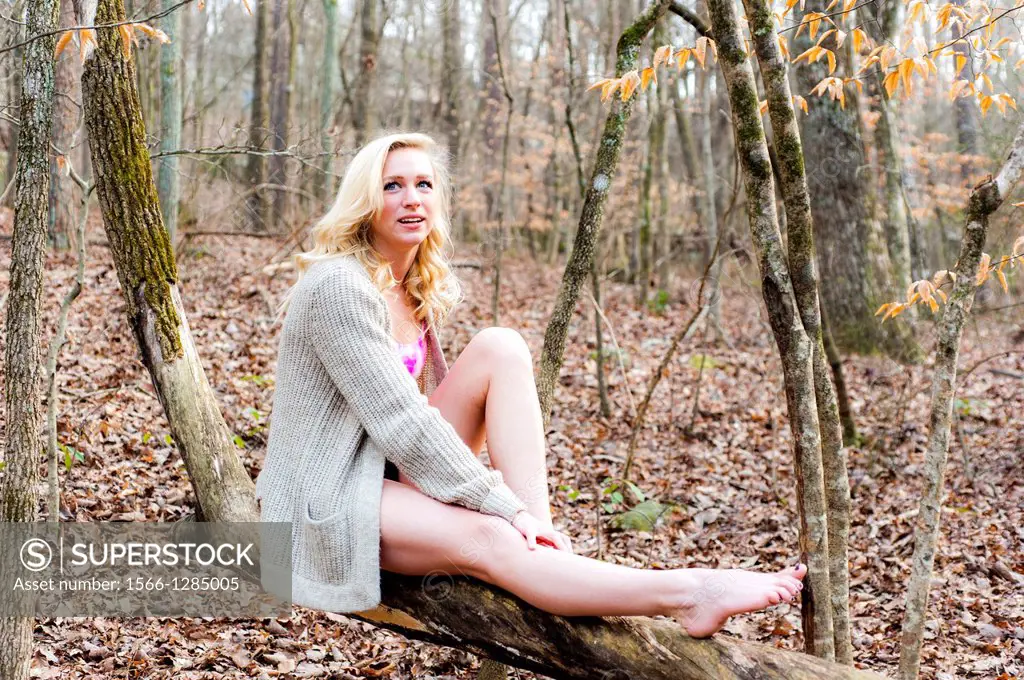 A partially nude 23 year old blond woman wrapped in a sweater sitting a tree branch in a forest setting.