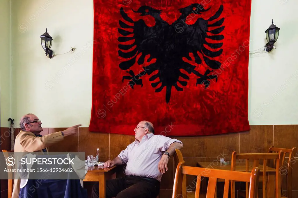 Men talking in a coffehouse by huge Albanian flag. Tirana, Albania.