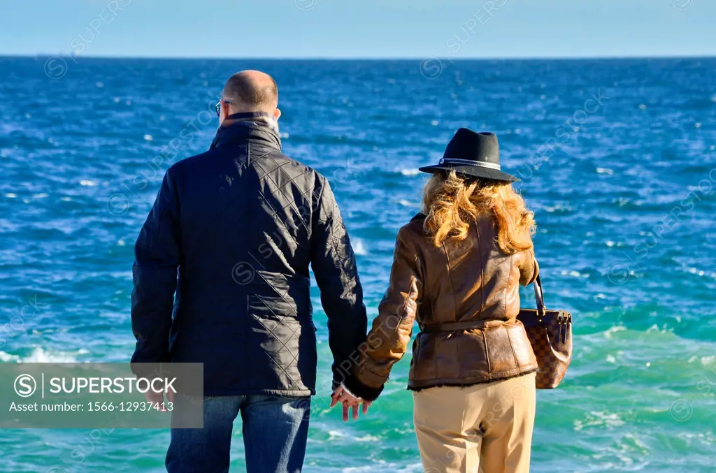 Adult couple watching the sea a sunny day. They are back and holding hands. They are dressing winter clothes.