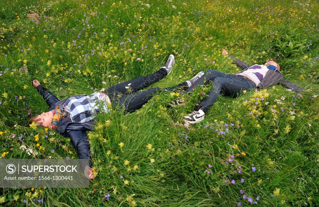 two young women laying with open arms, resting in the grass full of wildflowers, Saas Fee, Switzerland, canton Valais, caton Wallis