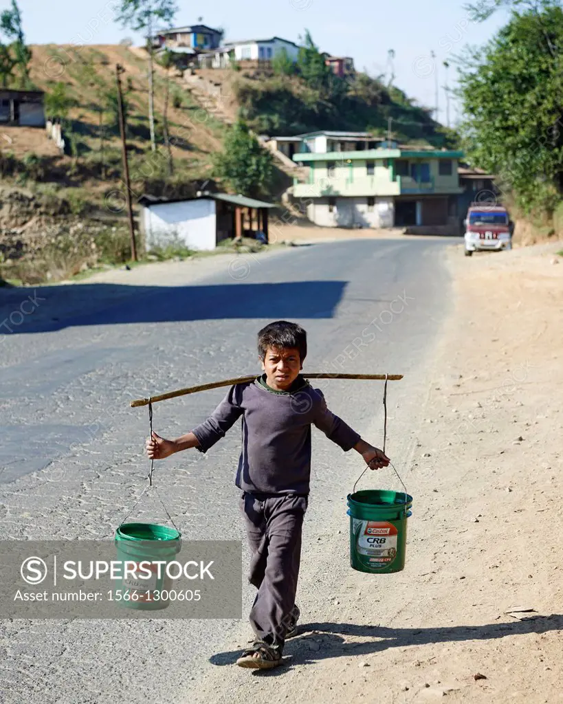 A young boy balances two green buckets while walking on the side of a road to a house.