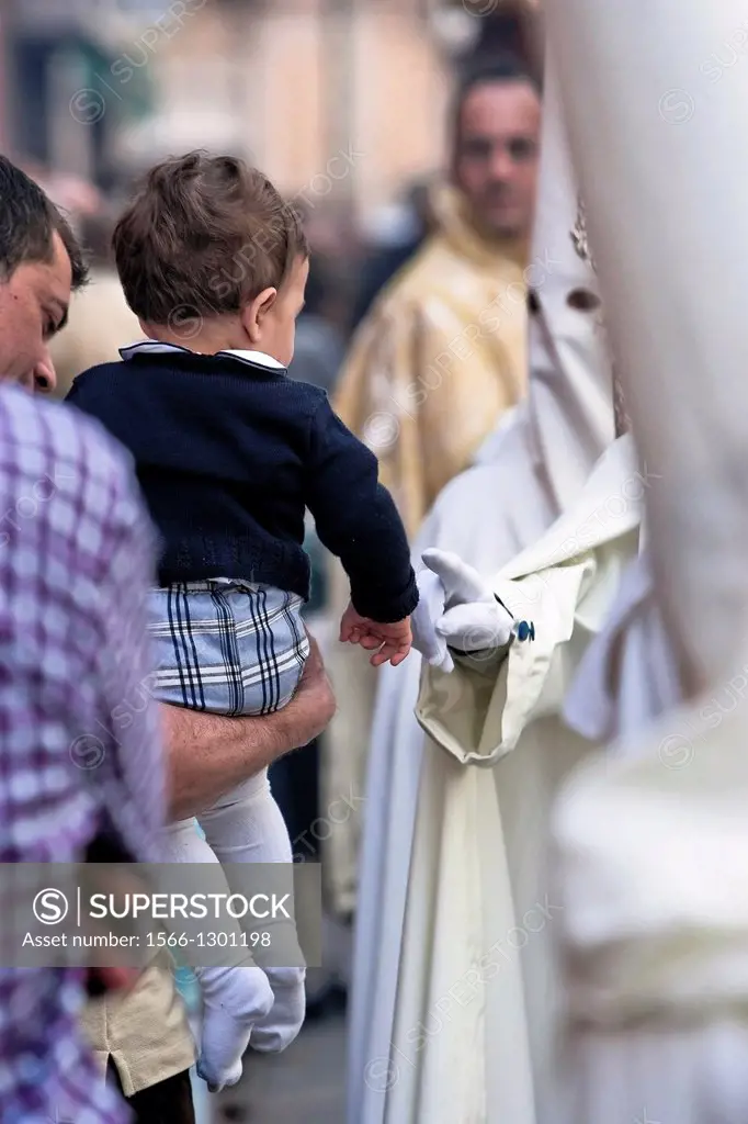penitent touches the finger of the hand of a child during a Holy week procession, Linares, Jaen province, Andalucia, Spain.