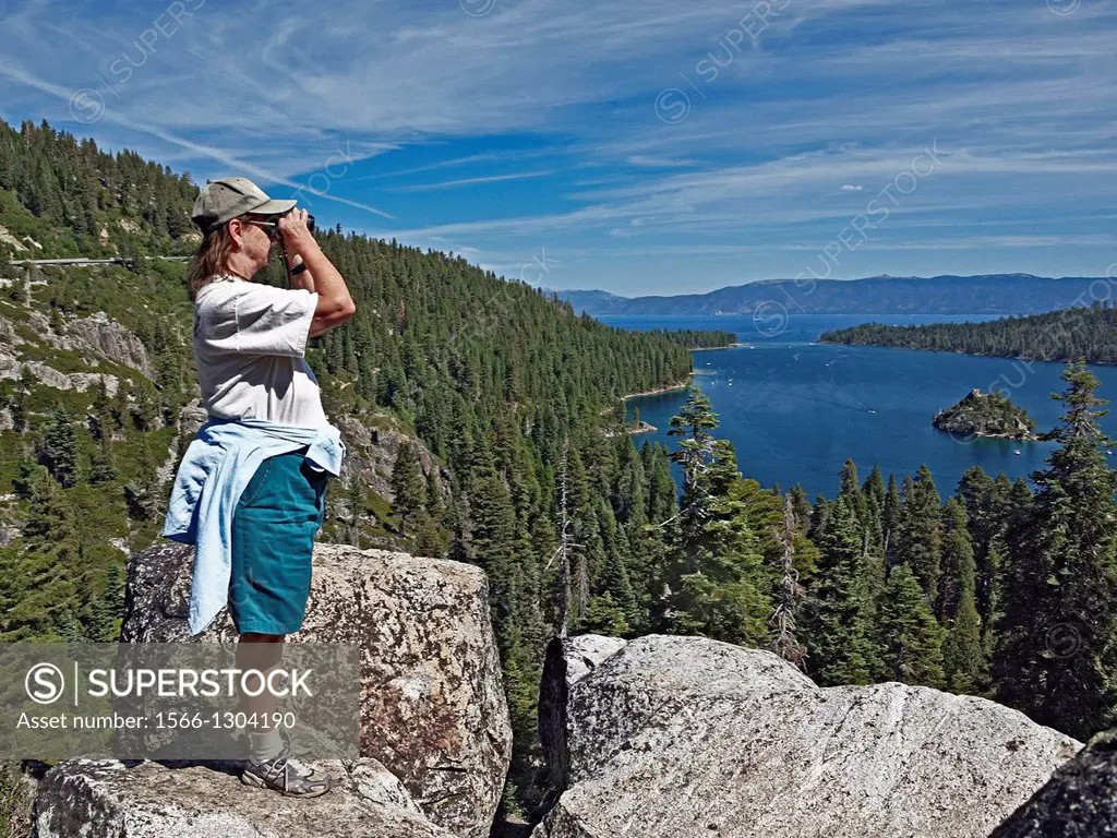 Woman standing on large rocks looking through binoculars at Lake Tahoe.