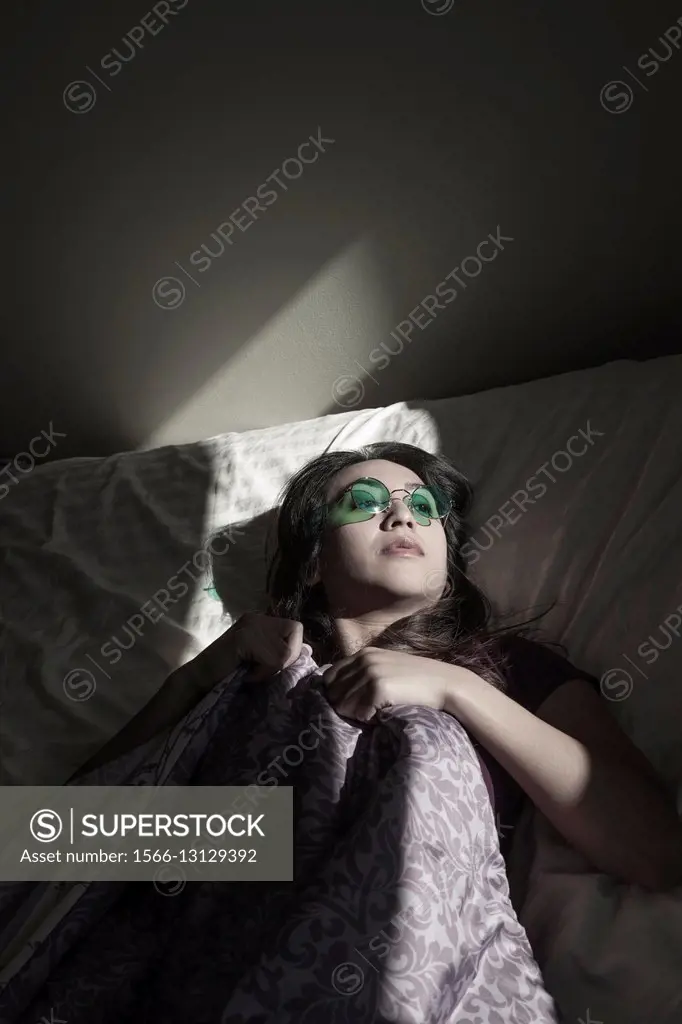 Young Asian woman lying on her bed, wearing colored glasses.