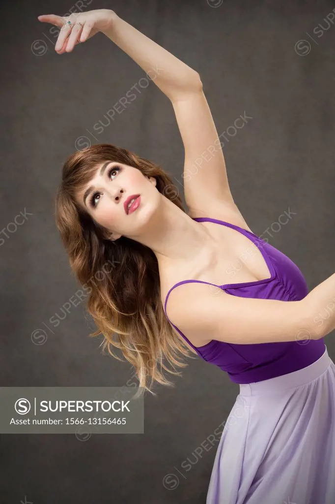 Attractive young woman dancer turning in purple leotard and lavender dress, from waist up on gray background.