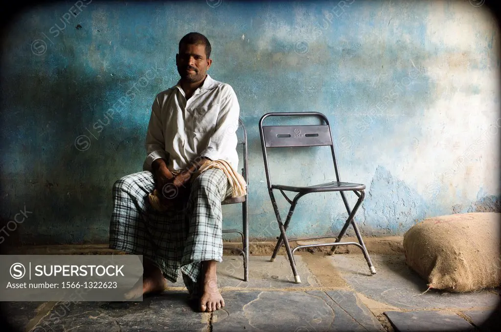 Portrait of one man sitting in a chair next to a sack on the ground, with blue background wall in a warehouse Mundgod, Karnataka, India, Asia