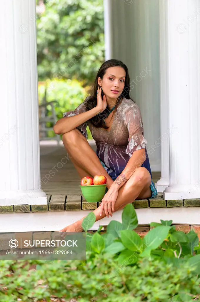 A 27 year old brunette woman wearing a short summer dress sitting on a porch with a bowl of apples.