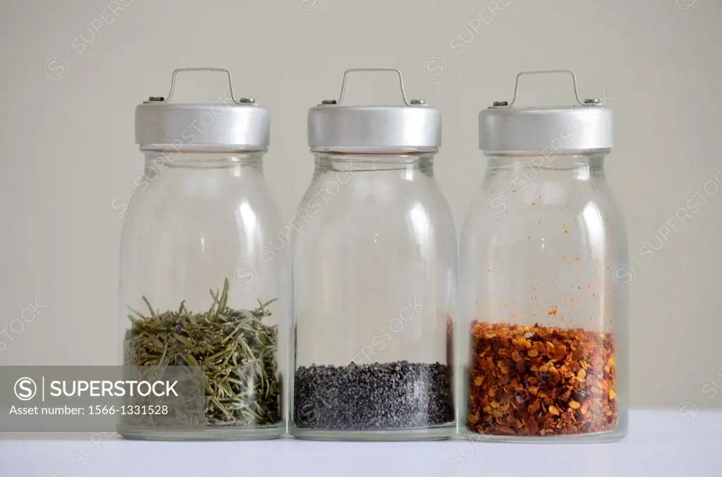 Glass jars with spices. From left to right: rosemary, poppy seed, chilli. Gray background.