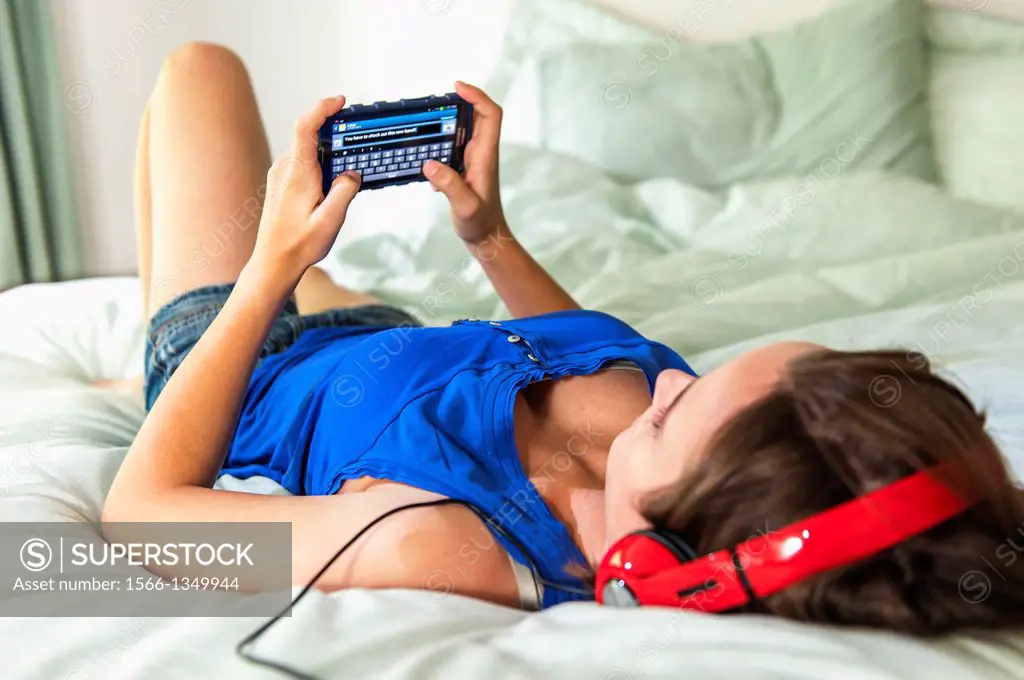 An 18 year old girl lying on her bed texting and weaing head phones.