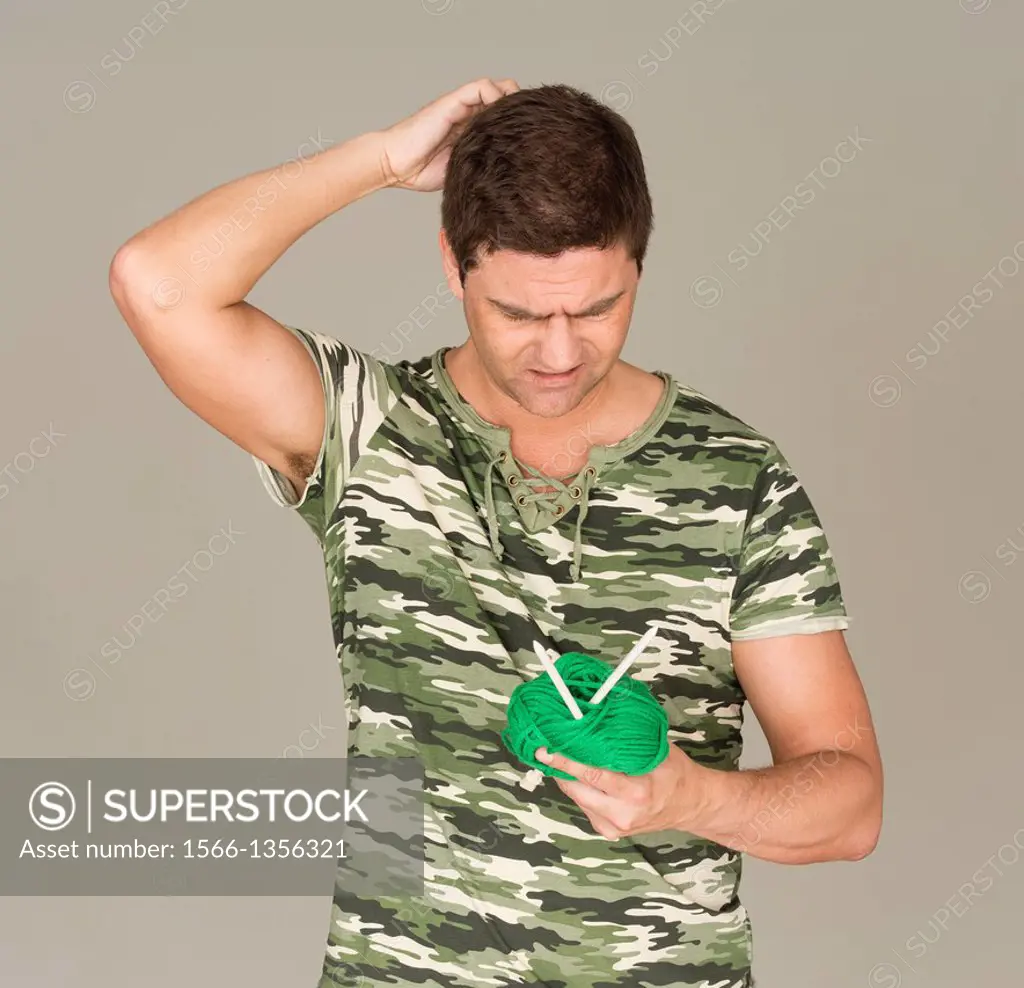 Man with camouflage t-shirt looking puzzled at a ball of yarn and knitting needles.