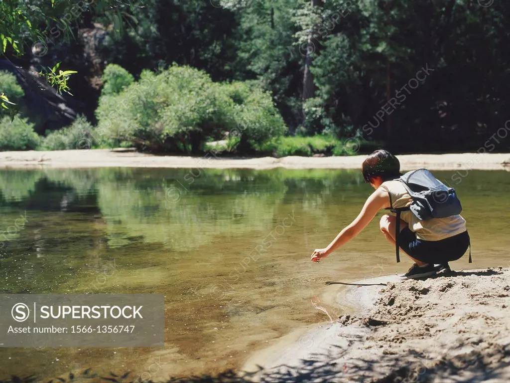 A young woman with a backpack dips an object in the river in Yosemite National Park.