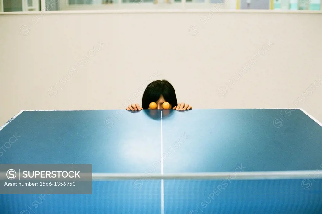 A woman poses with two orange ping pong balls as eyes at the edge of a ping pong table.