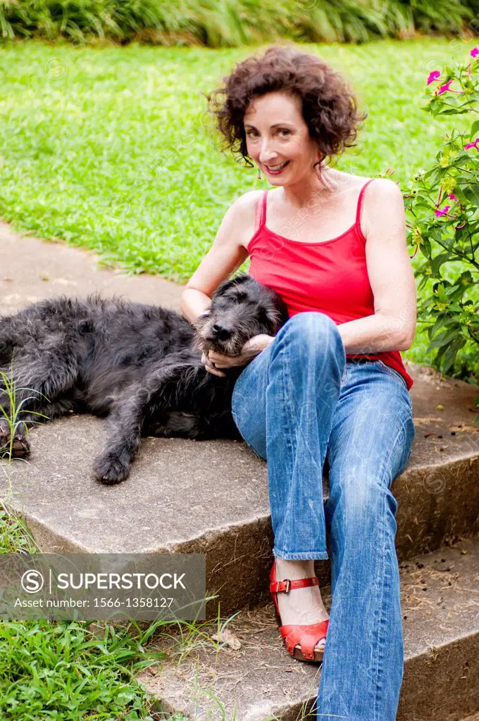 A 51 year old brunette woman wearing jeans sitting on steps in front of her house playing with her dog.