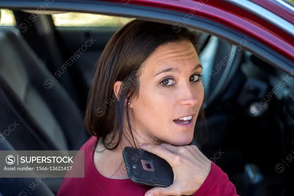 A 36 year old brunette woman in the passenger seat of a car, holding a cell phone and looking up at the camera.