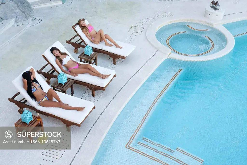 Three young attractive hispanic women using loungers for tanning next to swimming pool.