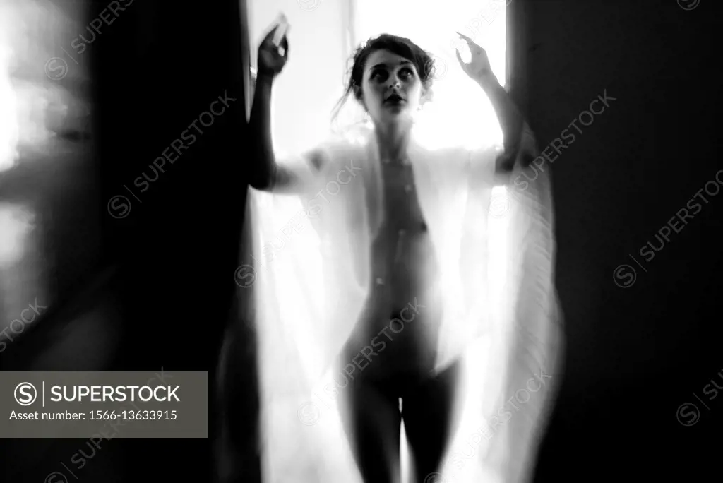 A nude and silhouetted young woman standing in a door way with her arms raised, black and white.