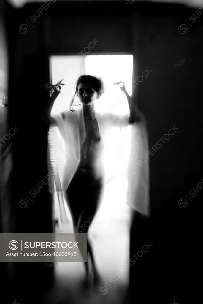 A nude and silhouetted young woman standing in a door way with her arms raised, black and white.