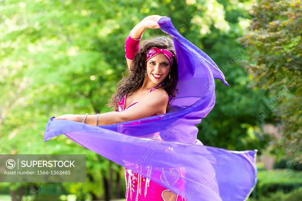A 23 year old brunette woman in costume belly dancing outdoors.