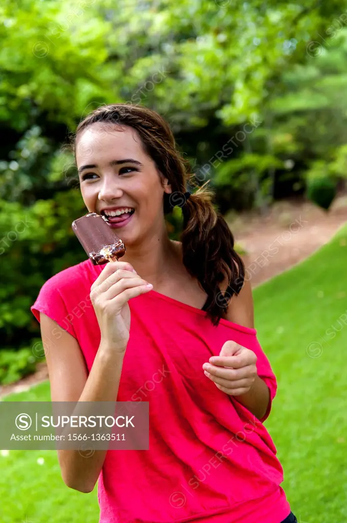 A 14 year old brunette girl eating a chocolate ice cream bar in a park setting.