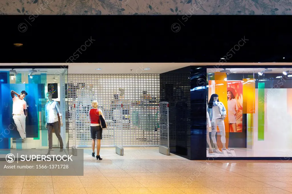 Woman standing at closed shop gate. Mannequins, dummy on store show-window. Retail shop entrance or exit. Facade of shop with display. Exterior.