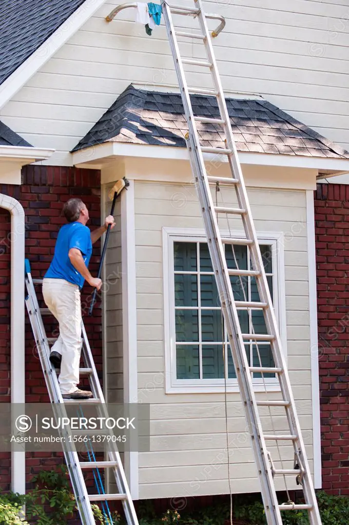 A 45 year old man on a ladder cleaning the wood siding of a house with a brush.
