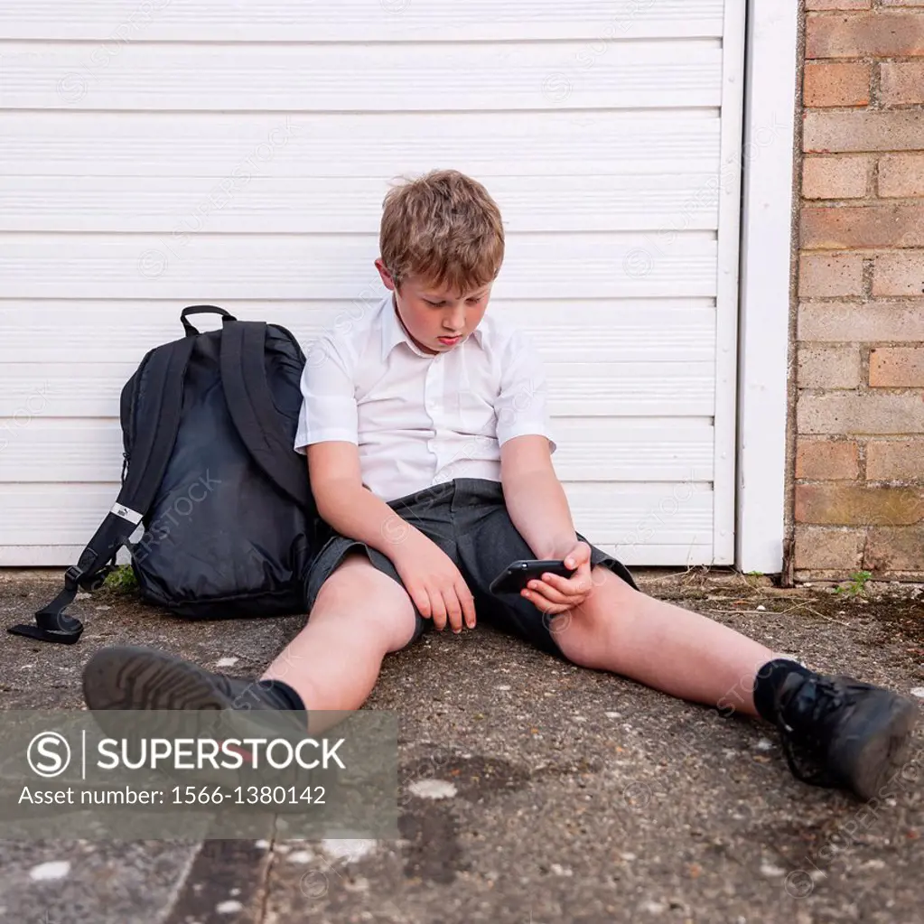 A boy of 10 looking sad and depressed in his school uniform as he looks at his mobile phone showing the effects of bullying by text or social networki...