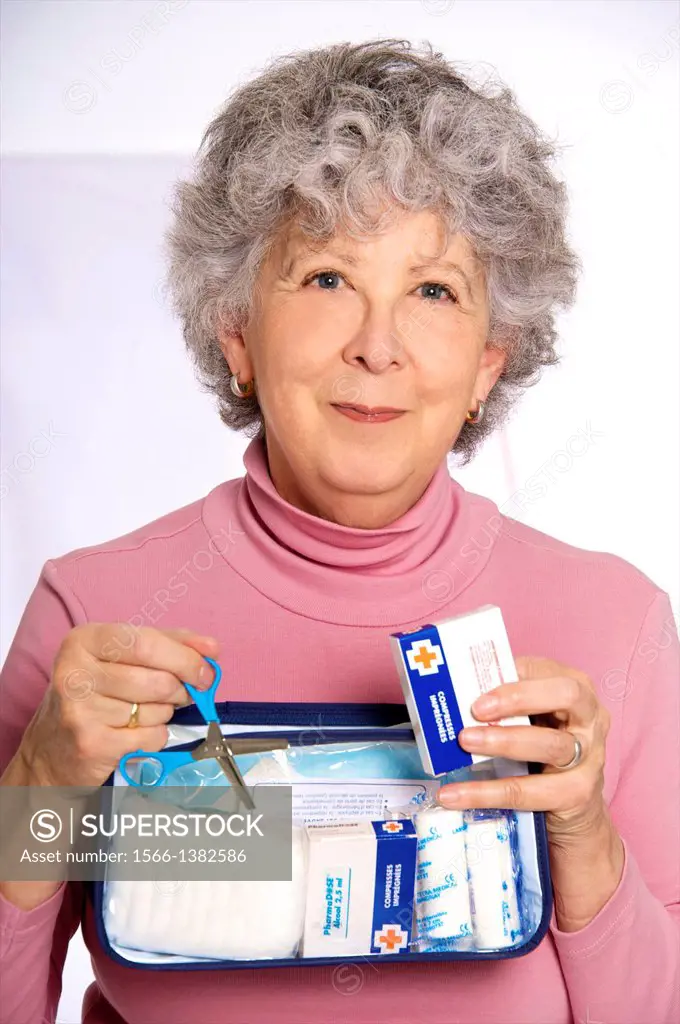 Senior woman, or senior nurse, in pink, holding transparent plastic first aid kit in her hands with blue scissors and dressings, at home or in surgery