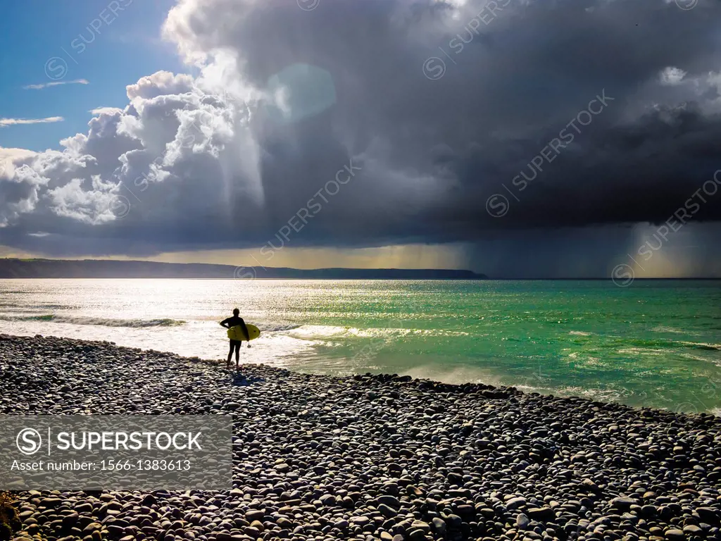 Surfer stood on a pebble beach looking out to sea with a rainstorm in the distance. Cornborough Range, Abbotsham, North Devon, England.