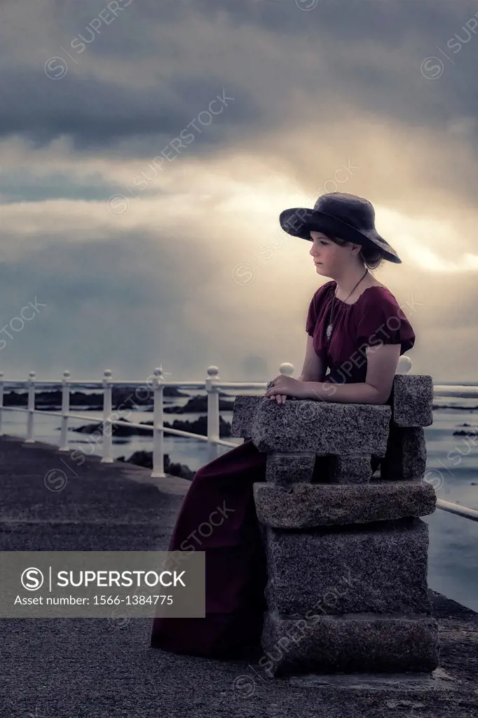 a sad girl with a black sunhat sitting on a stone bench at the sea.