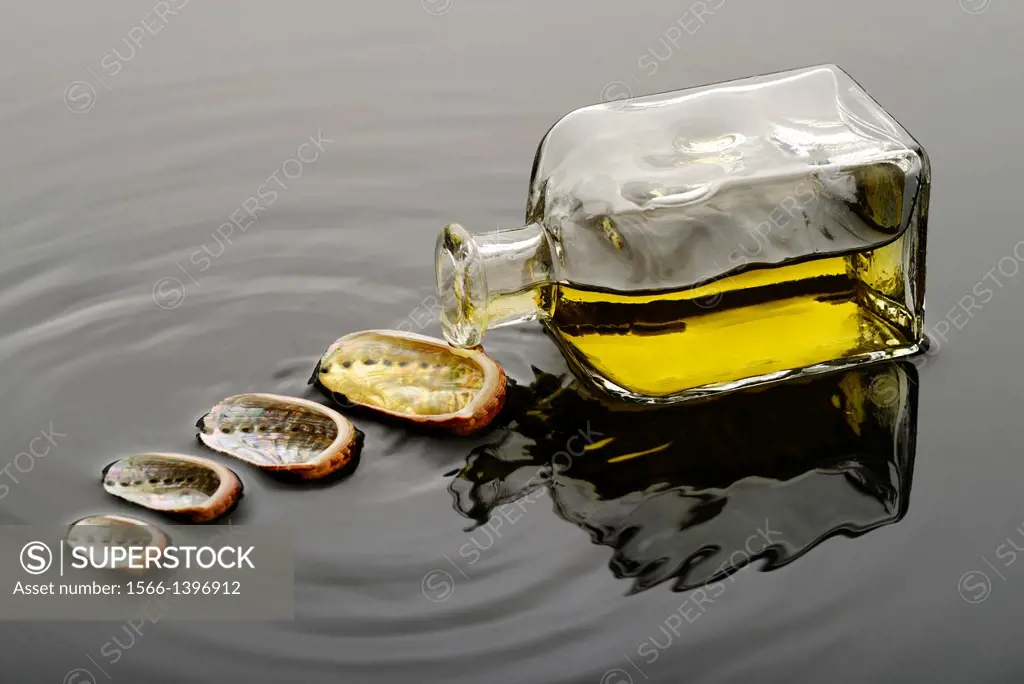 Yellow liquid, oil or perfume, spilling from a glass bottle over a seashell on water.