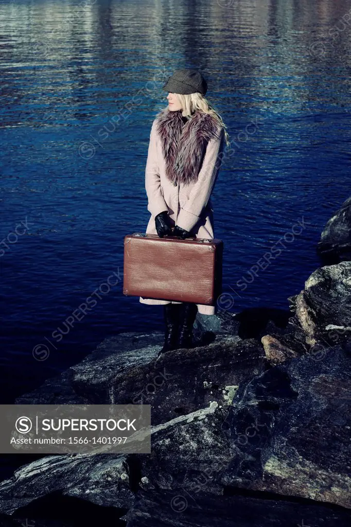 a girl in a pink coat is standing at the sea on stones with a suitcase.