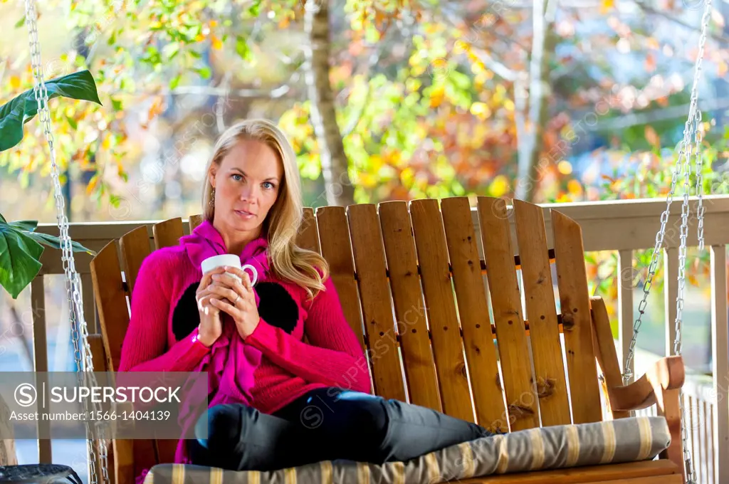 A portrait of a 39 year old blond woman wearing a sweater sitting on a porch swing holding a coffee cup, outdoors.