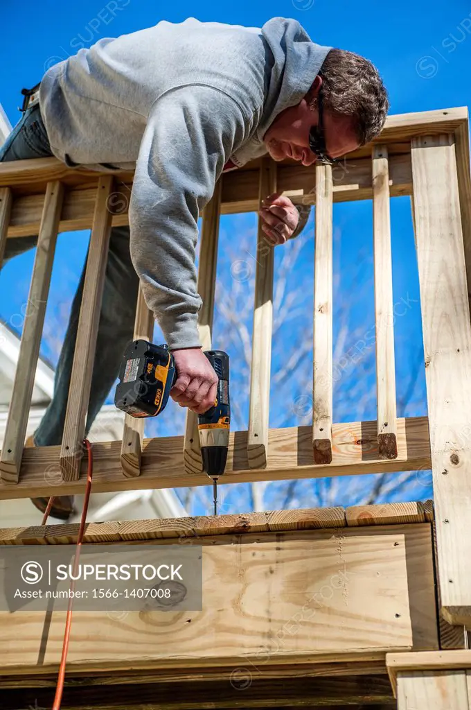 A man drills a screw into a piece of lumber on an outdoor deck.
