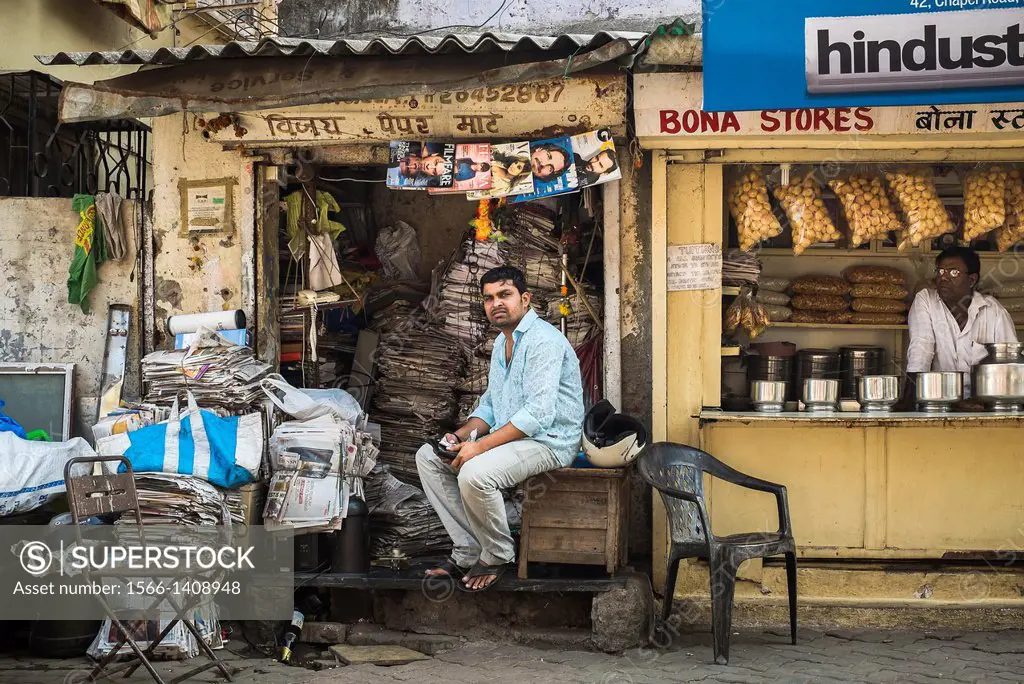 A young man sits clutching his tablet computer at his small shop where he recycles old newspapers and magazines; next to a shop selling supplies for I...