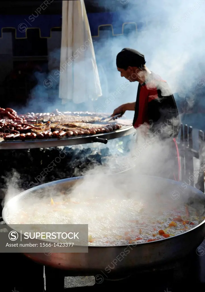 vegetables boiling in large pan and man preparing meats on giant barbecue in the annual All Saints Market in Concentaina, Alicante province, Spain.