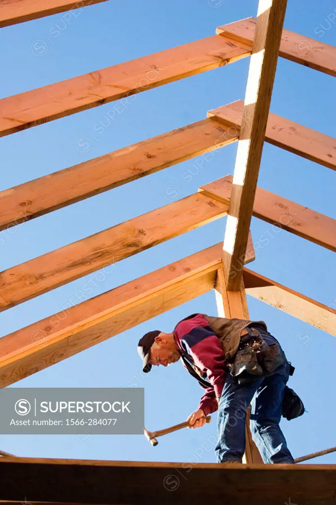 Carpenter hammering trimmer to center post while standing below the framing of the roof rafters at a construction site.