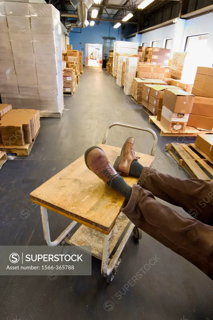 Man´s feet, wearing shoes, resting on a rolling cart in a factory´s storage area.