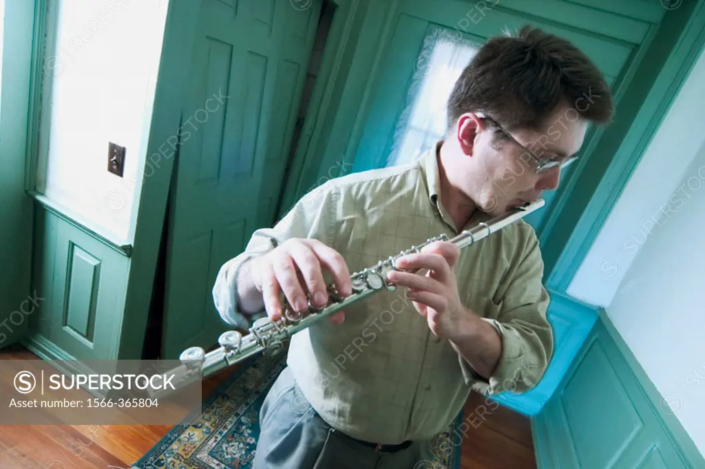 Young man, wearing glasses, playing a flute in the hallway of his house.