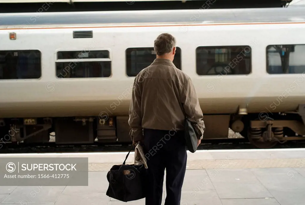 Rear view of a middle aged man holding a bag, waiting for train at Aberystwyth Railway station with Arriva wales train pulling into the station. Abery...