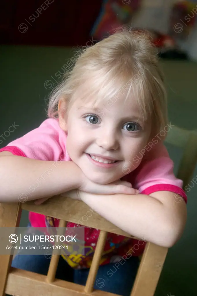 3 year old girl smiling into camera at nursery, leaning over the back of the chair onto her arms